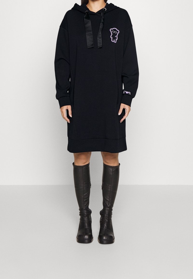 Schwarzes Kapuzenkleid mit langen Ärmeln, das ein lila Grafikdesign auf der Brust, satinierte Kordeln und ein seitliches Logo-Detail aufweist. Knielange schwarze Stiefel.