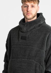 Pullover in pile di colore grigio scuro con colletto alto, parte frontale con mezza zip, tessuto testurizzato e una piccola patch nera con logo sul petto.