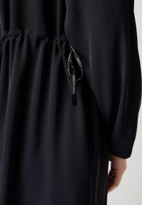 Gros plan sur un tissu de robe noire avec taille froncée et cordon noir étiqueté "Marc Cain Sport" près de la main d'une personne.
