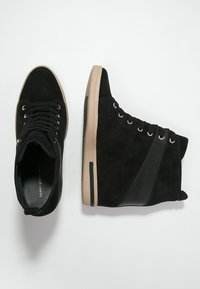 Svarta mocka hög-top sneakers med en tan gummisula, featuring snörning framtill, sidodekorer i läder och cirkulära perforeringar för ventilation.