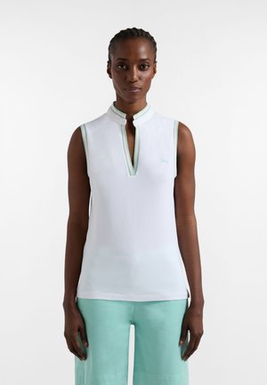 Kobieta w białym topie bez rękawów z zielonym wykończeniem i jasnozielonych spodniach, stojąca na tle jednolitego białego tła.