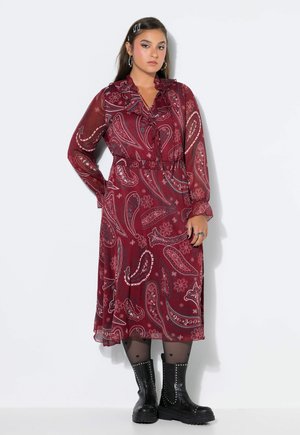 Mujer joven con un vestido largo rojo de paisley con volantes, medias negras transparentes con patrones de corazones y botas negras con tachuelas.