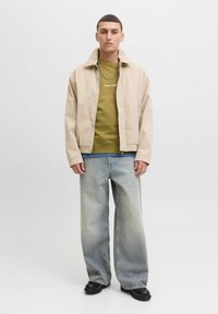 Giubbotto beige chiaro con design con colletto, felpa verde oliva sotto, e jeans larghi azzurri chiari. Scarpe nere completano il look.