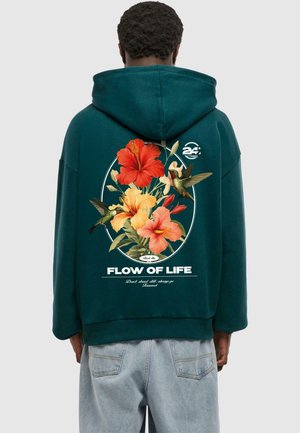 Mikina s kapucňou v tmavozelenej farbe s kvetinovou grafikou, ktorá zobrazuje červené a oranžové hibiscusy a kolibríky, text na spodnej časti.
