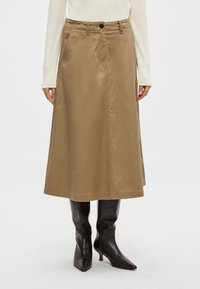 Brūna kokvilnas midi svārki ar taisnu griezumu, sānu kabatām un priekšpusē ar pogu aizdari, kombinēti ar melniem ādas zābakiem līdz ceļiem.