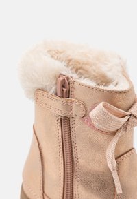 Botas cortas en oro rosa con un cierre de cremallera, forro de piel de conejo y un detalle decorativo de lazo. Presenta un acabado metálico texturizado y suela resistente.