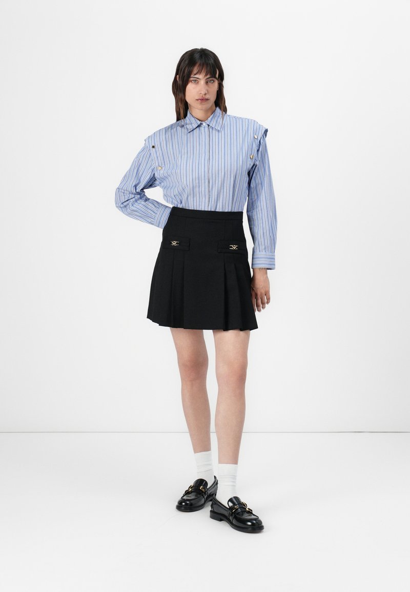 Blauw-wit gestreepte button-up shirt met open schouders, gecombineerd met een zwarte geplooide mini-rok met gouden accenten, en zwarte instapschoenen.
