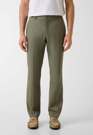 Uomo che indossa pantaloni verde oliva a gamba dritta, mocassini beige e una T-shirt bianca, in piedi davanti a uno sfondo semplice.