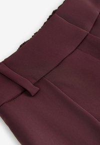Bordeaux stof, glat tekstur med plisseret talje og en snoet detalje. Nærbillede, der fremhæver syninger og design detaljer.