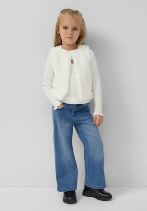 Jong meisje staat tegen een grijze muur, draagt een wit nepbonten vest, een shirt met lange mouwen, wijde blauwe jeans en zwarte laarzen.