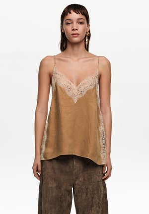 Mujer con una camisola de satén color canela con ribete de encaje beige y pantalones de ante marrón, de pie frente a un fondo blanco liso.