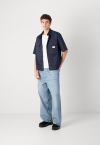 Marineblå kortermet skjorte med knappestemming foran, to brystlommer, kombinert med lyseblå vide jeans og svarte klumpete sko.