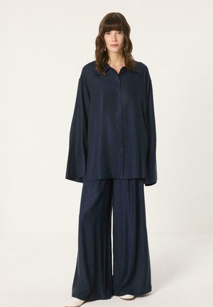 Kobieta ubrana w luźną granatową koszulę na guziki i szerokie spodnie, stoi na tle jednolitego, jasnego tła.