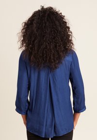 Camicia leggera a maniche lunghe di colore blu navy, con maniche a risvolto, vestibilità leggermente ampia e dettaglio plissettato nella parte posteriore. Tessuto morbido.