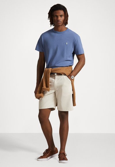 Mäns blå bomullströja, krämfärgade shorts, brun tröja knuten i midjan, bruna skor och en klocka. Enkelt mönster, avslappnad look.