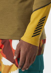 Olijfgroene longsleeve shirt met een gestructureerde gele mouw met zwarte diagonale strepen, gecombineerd met shorts met een multicolore patroon.