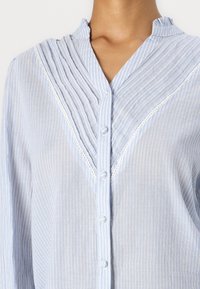 Camicia azzurro chiaro con bottoni, a strisce bianche verticali, design plissettato e pizzo lungo il collo. Tessuto morbido e leggero.
