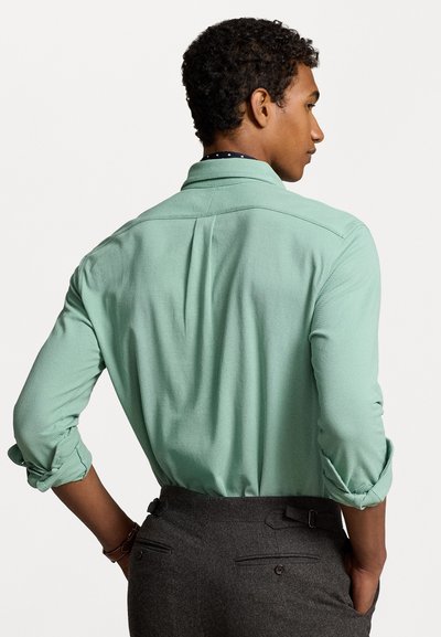 Camisa de manga comprida verde menta, tecido texturizado, mangas arregaçadas e colarinho pontudo, combinada com calças sob medida cinza escuro.