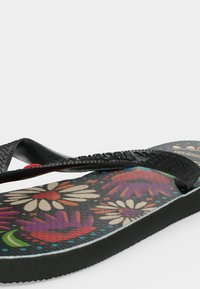 Tongs noires avec bride texturée et motifs floraux colorés en violet, rouge, blanc et vert sur la semelle sur fond uni.