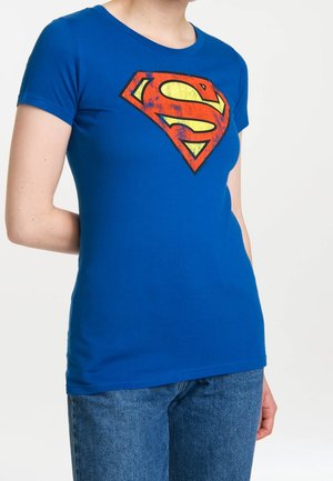 Personne portant un t-shirt bleu moulant avec un logo Superman rouge et jaune de style vintage, associé à un jean bleu.