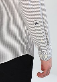 Camicia button-up a righe in bianco e blu navy, caratterizzata da un piccolo dettaglio ricamato sulla manica e polsini abbottonati, con un orlo curvo.