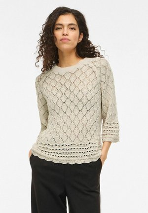 Femme aux cheveux bouclés portant un pull en tricot crème avec des motifs en losanges et des manches trois-quarts, associé à un pantalon noir.