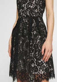 Robe noire en dentelle sans manches avec motif floral, taille cintrée et ourlet festonné, portée par une personne avec les bras détendus le long du corps.