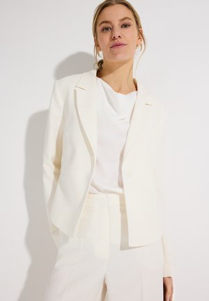 Vrouw met blond haar, gekleed in een off-white blazer, witte top en bijpassende broek, staand tegen een effen witte achtergrond.