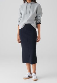 Sweat à capuche en coton gris avec cordon de serrage, associé à une jupe crayon bleu marine. Le mannequin porte des baskets blanches avec des accents bleus et gris.