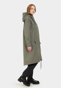 Impermeable largo de color verde oliva con capucha, cierre delantero con cremallera, cintura ajustable con cordón y detalles en negro. Llevado sobre pantalones y botas negros.