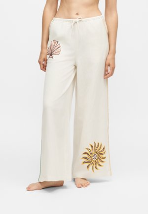 Femme portant un pantalon blanc ample avec un motif coquillage rose sur la cuisse gauche et un motif soleil jaune sur le mollet droit, pieds nus.