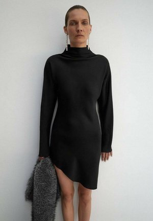 Robe noire à manches longues en tissu lisse, avec un col haut et un ourlet asymétrique. Le mannequin tient une étole en fourrure grise.