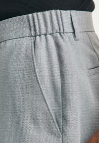 Pantaloni grigio chiaro su misura con tessuto strutturato, vita elastica e tasche laterali. Il design presenta motivi incrociati sottili.