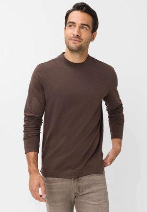 Uomo con capelli scuri che indossa una camicia marrone a maniche lunghe e pantaloni marrone chiaro, in piedi e che guarda leggermente verso l'alto di lato.
