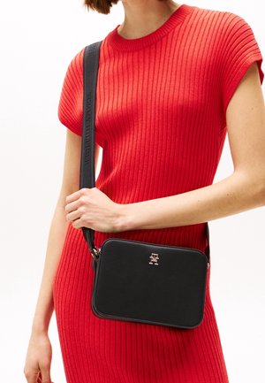 Schwarze rechteckige Umhängetasche mit glatter Textur, ausgestattet mit einem metallischen Logo und einem Stoffgurt. Das Modell trägt ein geripptes rotes Kleid.