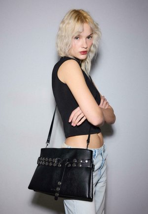 Plavuša u crnoj top bez rukava i svijetlim trapericama nosi crnu kožnu torbu s srebrnim zakovicama na ramenu.