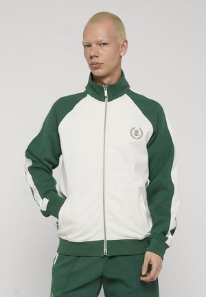 Ung person med kort blondt hår iført grøn og hvid lynlåstrøje og matchende bukser foran en neutral baggrund.