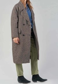 Cappotto doppiopetto in tessuto houndstooth marrone e beige, realizzato in lana testurizzata, con grandi bottoni e una vestibilità rilassata. Indossato sopra una camicia di jeans.