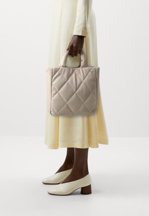 Person i cremefarvet kjole holder en beige quiltet håndtaske og bærer cremefarvede sko med lav hæl mod en ensartet lys baggrund.