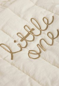 Quiltad filt i krämfärgad tyg med metallisk gyllene broderad text "lilla underverket". Mjuk textur med horisontell sömnadsmönster.