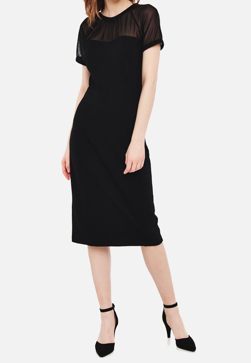 Femme portant une robe noire cintrée avec des manches courtes transparentes et des talons hauts noirs à bride cheville, debout devant un fond uni.