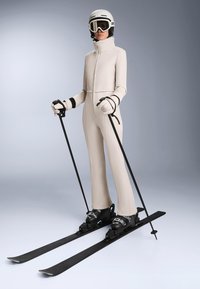 Kombinezon narciarski w kolorze kremowym, z suwakiem z przodu, wysokim kołnierzem i dopasowanym krojem. Czarna narciarska obuwie, kijki i gogle dopełniają strój.