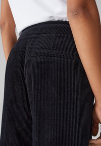 Black corduroy pants with prominent vertical ribbing, featuring a waistband and back pockets, captured from a side angle.