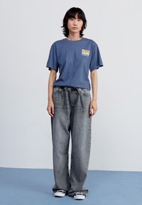 Blå bomull t-skjorte med et lite grafisk motiv på brystet, parret med løse, grå denim jeans og svarte joggesko. Enkelt og avslappet design.