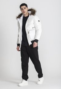 Giacca a vento bianca con cappuccio, con bordo in pelliccia, cerniere dorate e tasche laterali, abbinata a pantaloni neri e sneakers bianche.