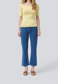 Gul tætsiddende top med korte ærmer, parret med mid-rise blå flared jeans og brune ruskind åben-tå mules. Enkel design uden mønstre.