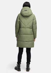 Padded green parka z kapturem, z chevronowym wzorem na plecach i prostym krojem. Noszony na czarnych spodniach i butach.
