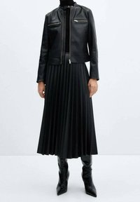 Veste en cuir noir avec deux poches zippées, associée à une jupe plissée noire. La tenue comprend des bottes noires montantes.