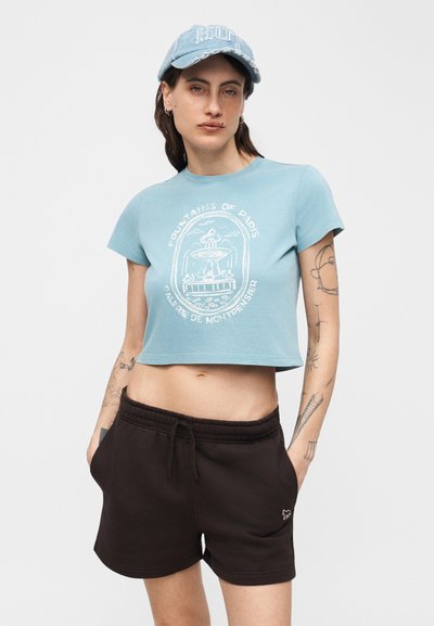 Γυναίκα που φοράει μπλε κοντομάνικη μπλούζα με στάμπα σιντριβανιού, μαύρα σορτς με κορδόνι, και ανοιχτό μπλε καπέλο μπέιζμπολ, στέκεται με τα χέρια στις τσέπες.