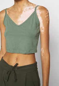 Donna con vitiligine sulla spalla e sul braccio, che indossa un top corto verde chiaro e pantaloni verde scuro con cordino.